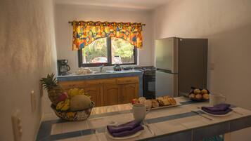 A kitchen with a refrigerator, stove, and window with curtains. There is a fruit bowl on the counter and a plate of pastries.