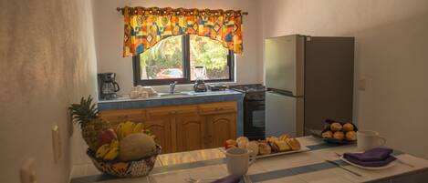 A kitchen with a refrigerator, stove, and window with curtains. There is a fruit bowl on the counter and a plate of pastries.