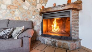 A stone fireplace with a fire burning inside, a grey sofa with patterned cushions, and a tiled floor.