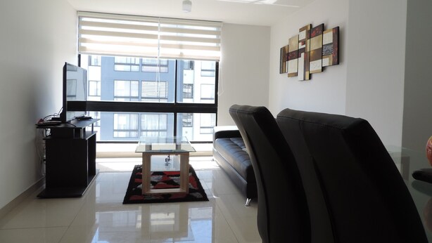 Una sala de estar moderna con un sofá, una mesa de centro y un televisor sobre un mueble. Se ve un ventanal con persianas y una obra de arte en la pared.
