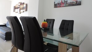 Un comedor moderno con una mesa de vidrio y sillas negras, un frutero sobre la mesa y un cuadro de un paisaje urbano en la pared.