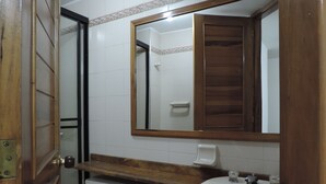 Un baño con puerta de madera, baldosas blancas, un espejo, un lavamanos y un inodoro.