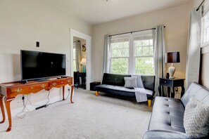 Un salon avec un téléviseur posé sur un meuble en bois, un canapé noir, un canapé gris et une fenêtre avec des rideaux.
