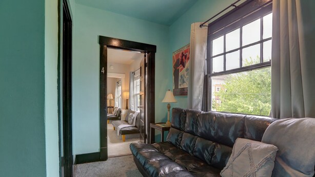 A room with a dark leather sofa, a small table with a lamp, and a window with curtains. A door leads to another room with a chair and a lamp. - Your own historic estate in the heart of Old Louisville! NEAR IT All!! (Louisville)