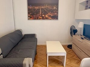 Un pequeño espacio de sala con un sofá gris, una mesa blanca y vista a la ciudad.