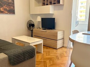 Un espacio habitable compacto con sofá, una mesita, televisión sobre un mueble de madera y una zona de comedor con una silla.