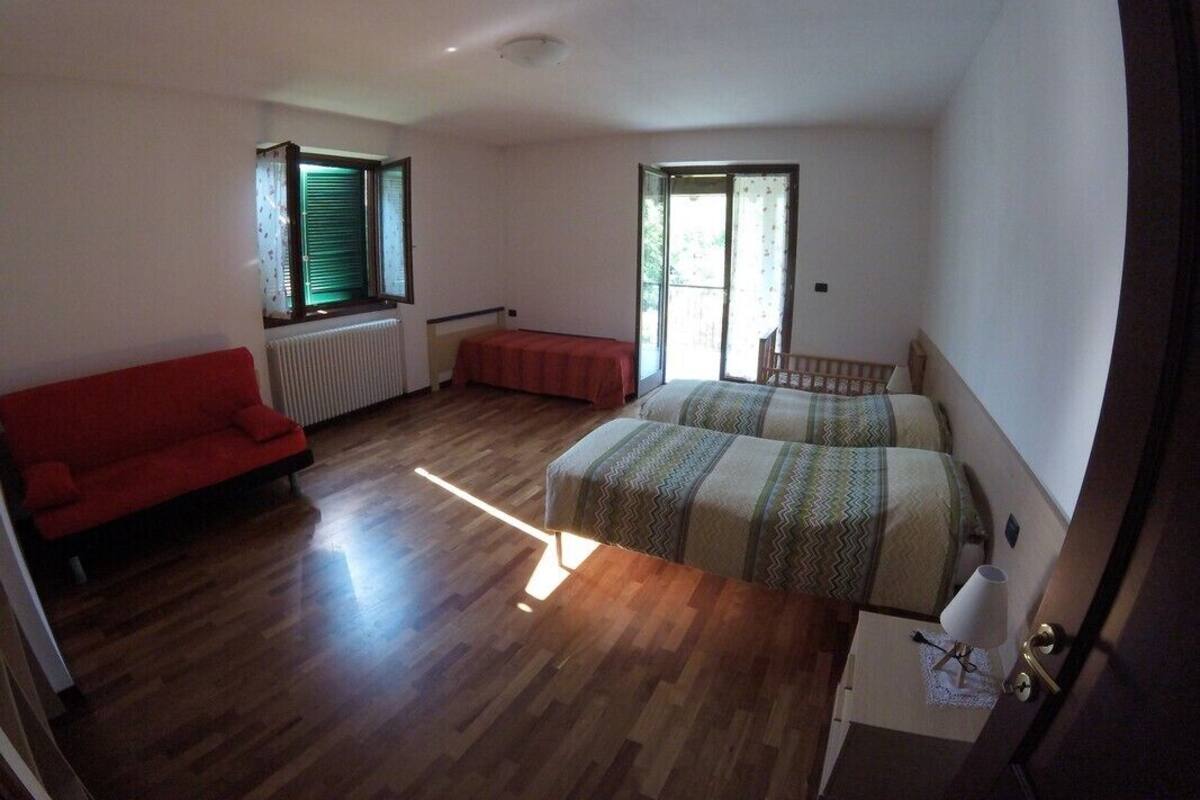 A bedroom with a wooden floor, a bed with a striped bedspread, a red sofa, and a window with blinds.