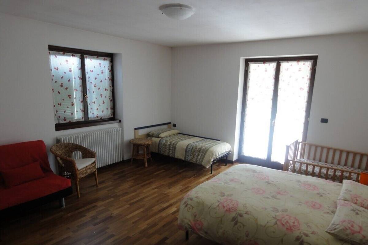 A bedroom with a wooden floor, a red sofa, a bed with floral bedding, a small table, and two windows with curtains.