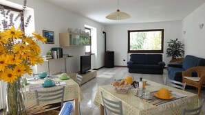 A living room with a sofa, a dining table with chairs, a TV, and a window with a view of greenery. - B&B Carunei (Poggiridenti)
