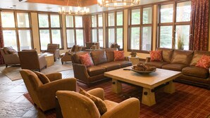 A spacious living room with leather sofas, a wooden coffee table, and a chandelier. Large windows provide natural light and views of the outdoors. - TRUE Ski-In Ski-Out Luxury 2 bd. 2 bth -Riverbend Lodge -Perfect location! (Breckenridge)