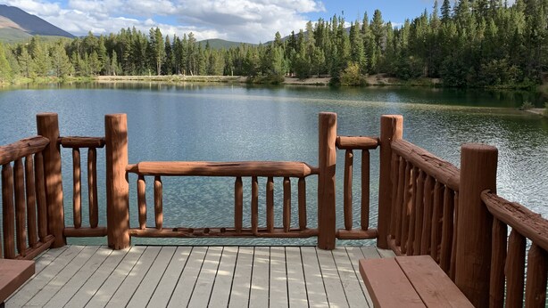 A wooden deck with a railing overlooking a calm lake, surrounded by dense forest and mountains. - TRUE Ski-In Ski-Out Luxury 2 bd. 2 bth -Riverbend Lodge -Perfect location! (Breckenridge)