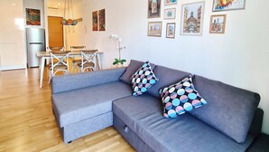 Un salon moderne avec un canapé d’angle gris, une table à manger avec des chaises et, au fond, un coin cuisine.