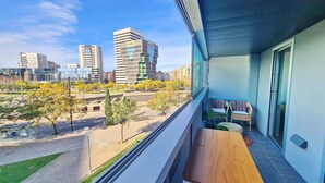 Un balcon avec une table en bois et deux chaises en osier, donnant sur un paysage urbain composé d’immeubles modernes et d’un parc.