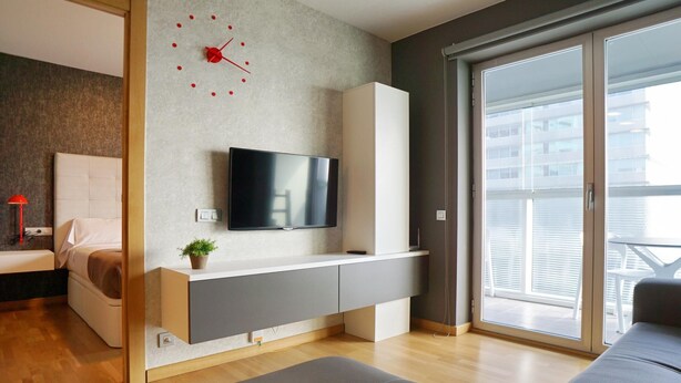 A modern living room with a flat-screen TV mounted on a wall, a sofa, a bed with a headboard, a small plant, and a sliding glass door leading to an outdoor area. - Apartment in Europe Square (Hospitalet de Llobregat)
