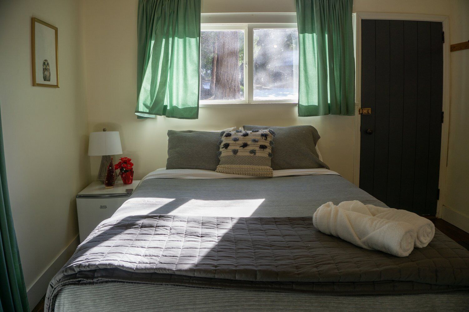 Een net opgemaakt bed met een grijze deken, een wit kussen en een gevouwen witte handdoek. Er staat een klein tafeltje met een lamp en een rode vaas met bloemen. Groene gordijnen bedekken de ramen, en een donkere deur is aan de rechterkant te zien.