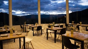 Un restaurant avec des tables et des chaises disposées pour accueillir les clients, de grandes fenêtres offrant une vue sur le ciel et les montagnes lointaines.