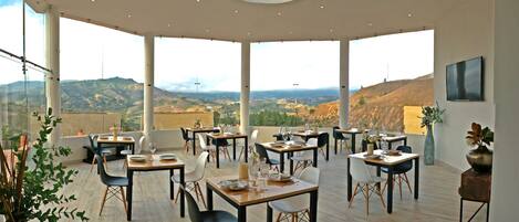 A spacious dining area with multiple tables and chairs, large windows offering a scenic view of the hills, and a modern ceiling design.