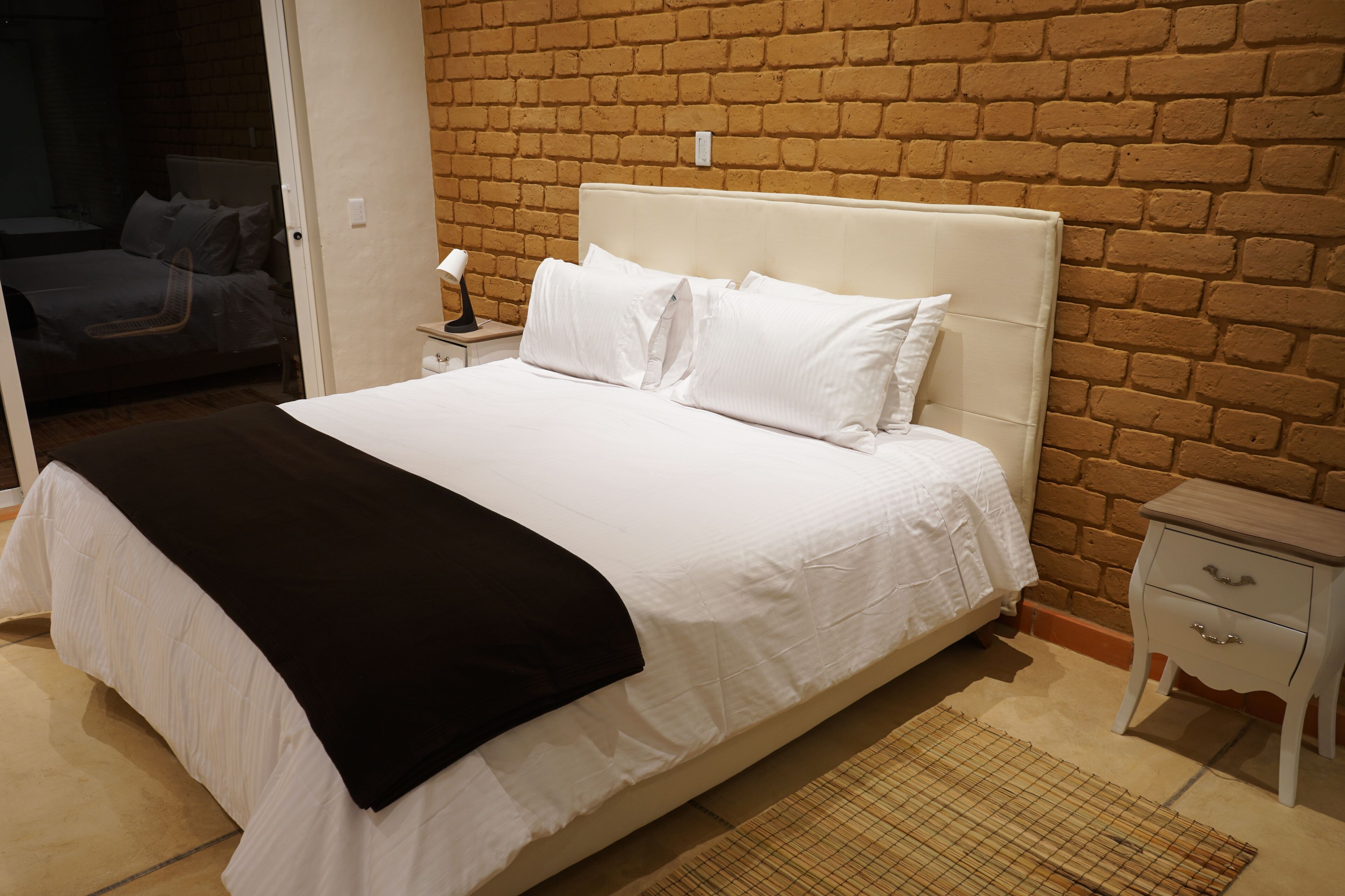 Una cama bien hecha con ropa de cama blanca y una manta de color negro, una mesita de noche con una lámpara y una pared de ladrillo al fondo.