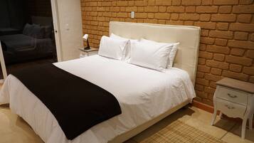 Una cama bien hecha con ropa de cama blanca y una manta de color negro, una mesita de noche con una lámpara y una pared de ladrillo al fondo.