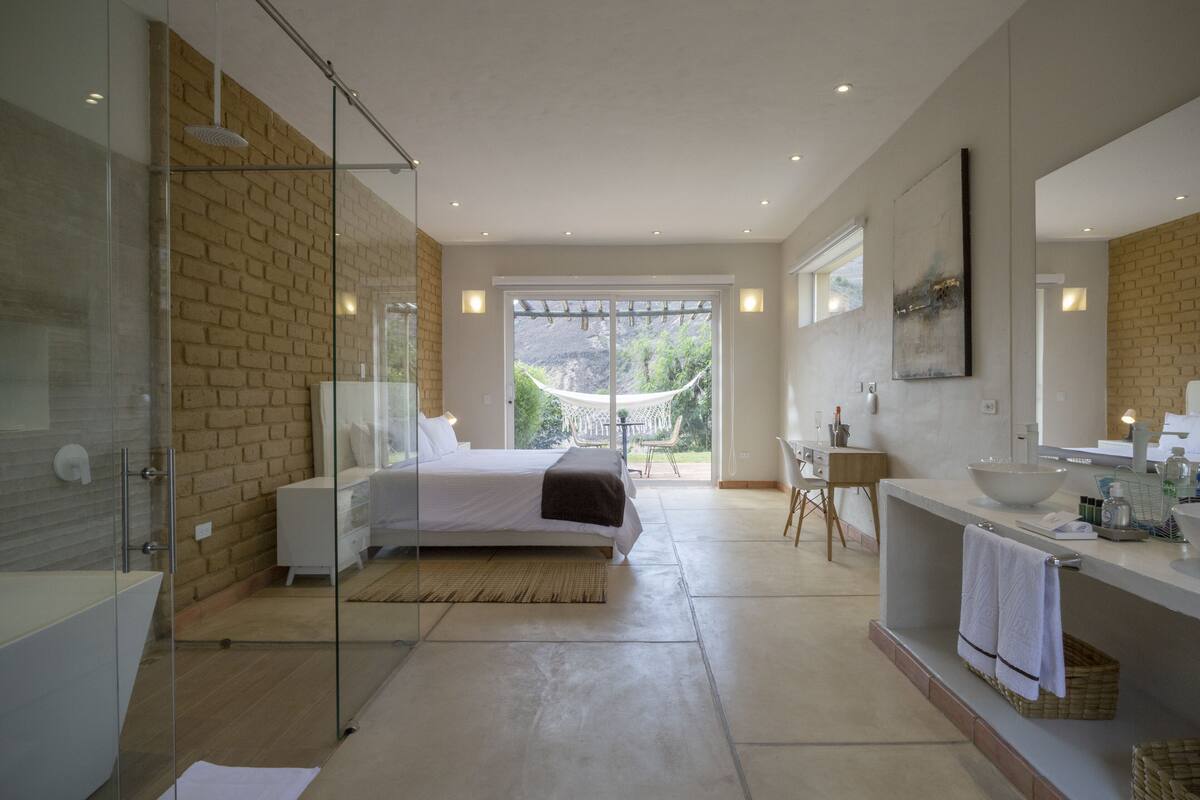 A modern bedroom with a large bed, a glass shower enclosure, a sitting area with a view of the outdoors, and a bathroom with a large mirror and sink.