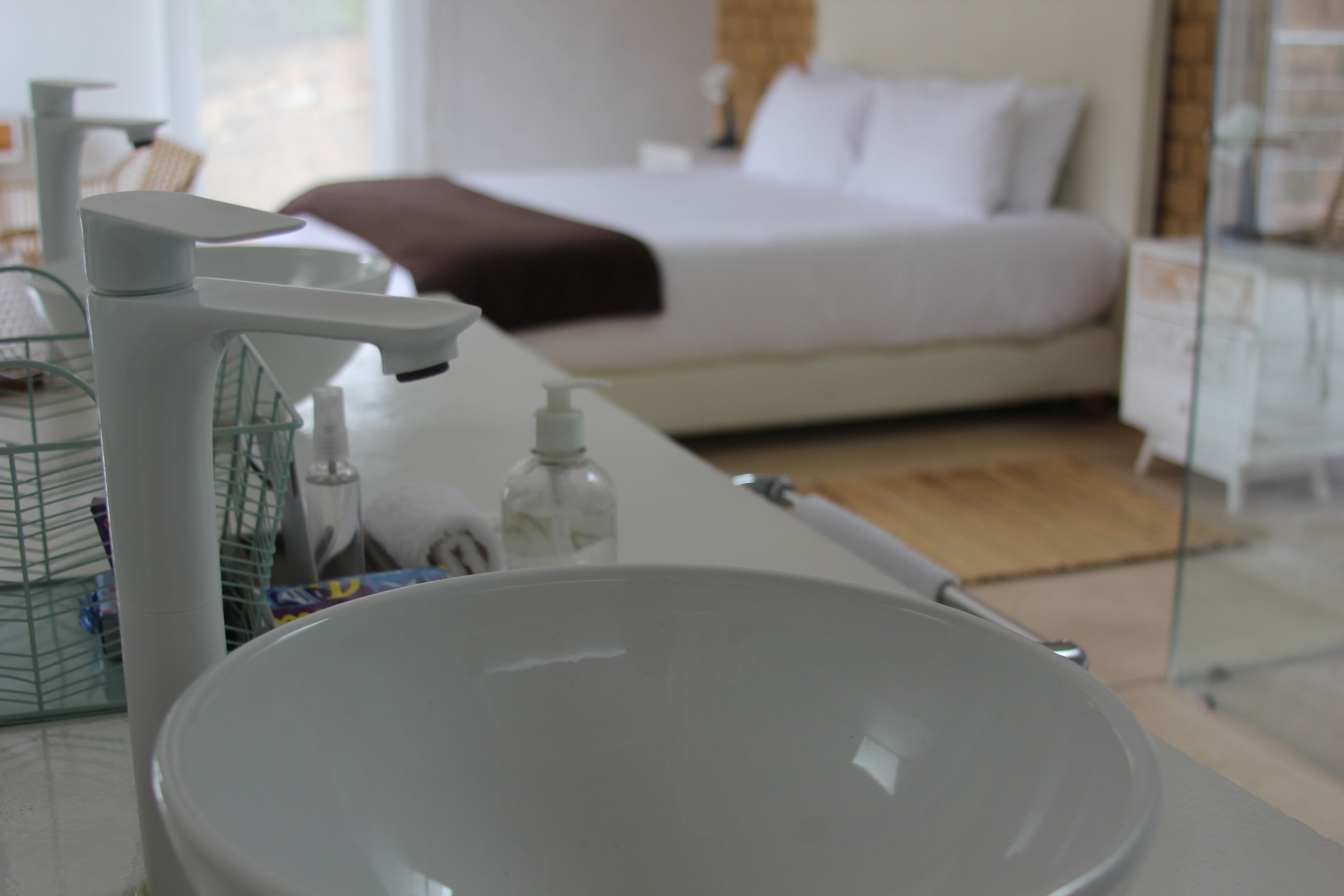 Une salle de bain moderne avec un grand lavabo blanc, une douche en verre et un lit en arrière-plan.