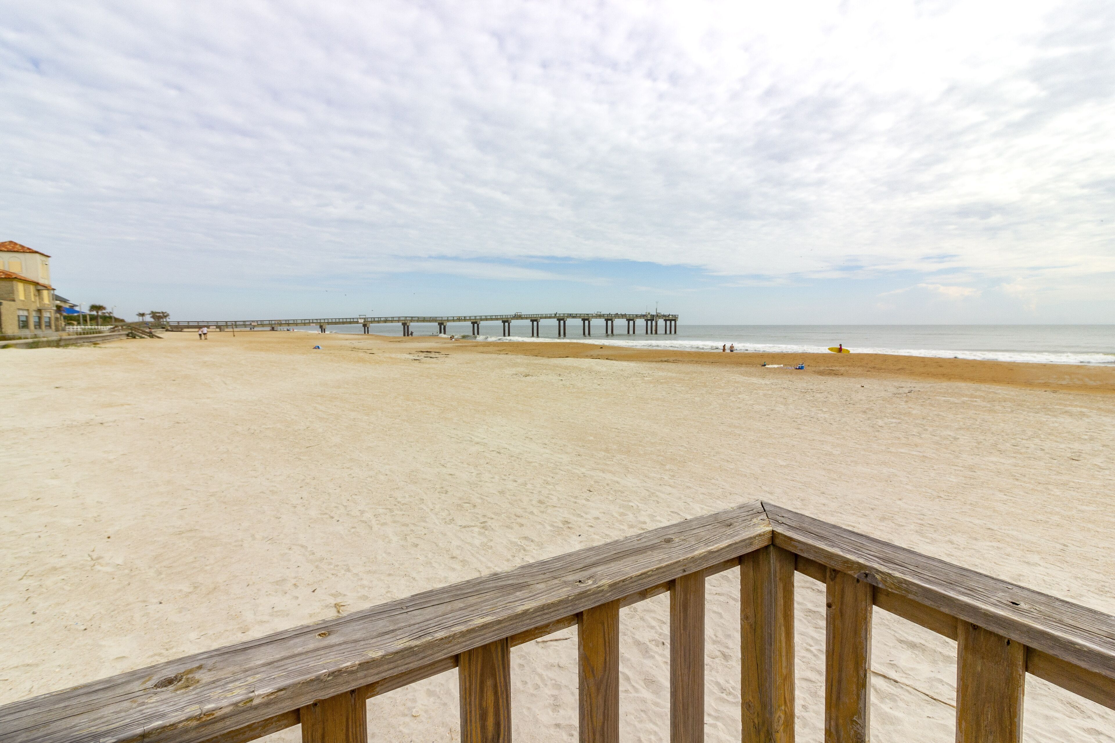 A beach with a wooden pier extending into the sea, a sandy shore, and a building on the left.