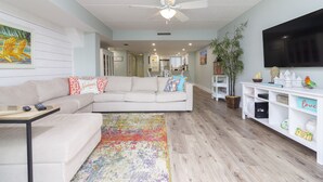 A living room with a sofa, a coffee table, a colorful rug, a ceiling fan, and a TV on the wall.