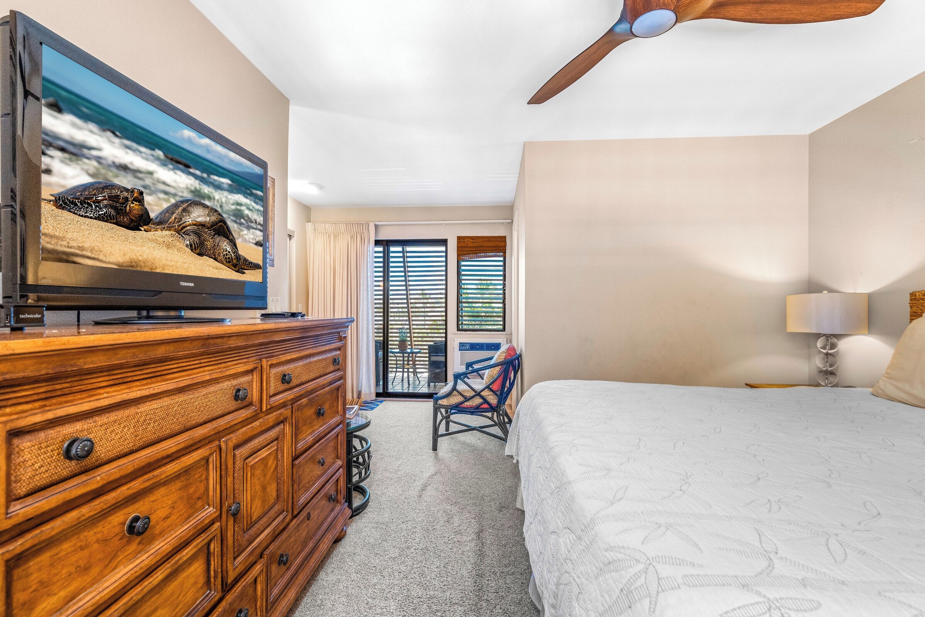 Une chambre à coucher comprenant un lit, une commode, un téléviseur diffusant une scène de plage, un ventilateur de plafond et une vue sur l’océan par une porte coulissante.