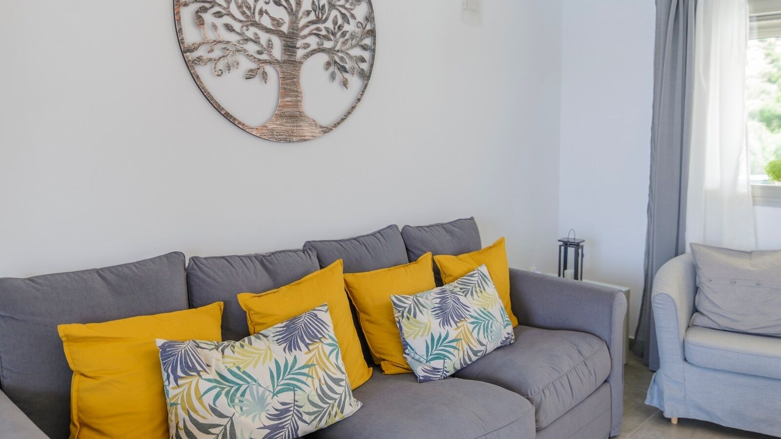 A grey sofa with yellow and leaf-patterned cushions, a wall-mounted tree decoration, and a grey armchair.