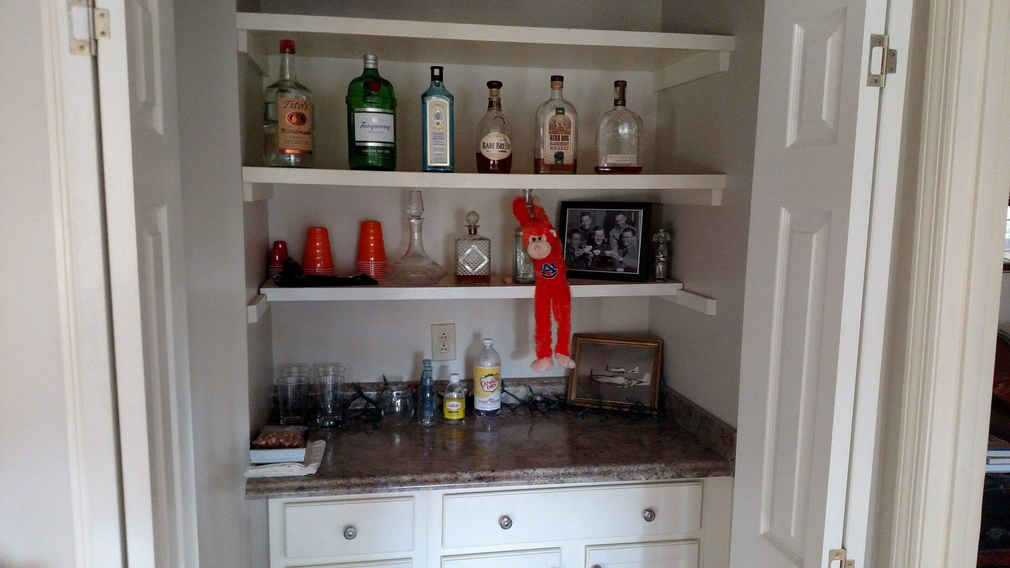 A kitchen with a countertop, shelves with liquor bottles, red plastic cups, and a framed photo.