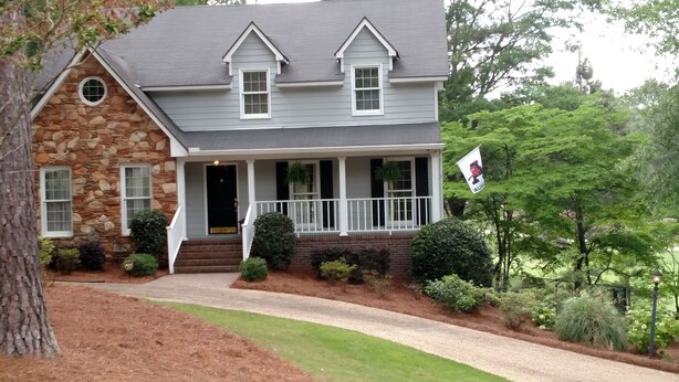 A two-story house with a stone facade, a front porch with white railings, and a flag with a cartoon character. - Auburn Gameday Retreat (Opelika)