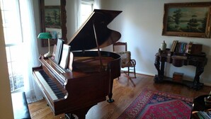 A grand piano in a well-lit room with a wooden floor, a chair, a table with books, and a painting on the wall. - Auburn Gameday Retreat (Opelika)