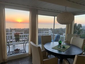 A dining area with a table and chairs, a hanging light fixture, and a view of the sunset over the cityscape. - Now booking Fall/Winter- Charming Beach View Cottage (Laguna Beach)