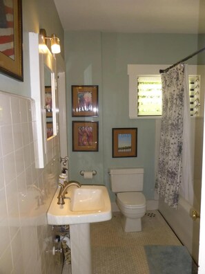 A bathroom with a sink, toilet, and shower. There are framed pictures on the wall and a window with blinds. - Now booking Fall/Winter- Charming Beach View Cottage (Laguna Beach)