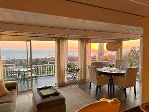 A balcony with a dining table and chairs, a sofa, and a view of the ocean at sunset. - Now booking Fall/Winter- Charming Beach View Cottage (Laguna Beach)