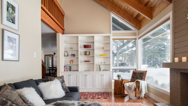 Una sala de estar acogedora con chimenea, vigas de madera, una estantería y un ventanal con vistas a árboles cubiertos de nieve.