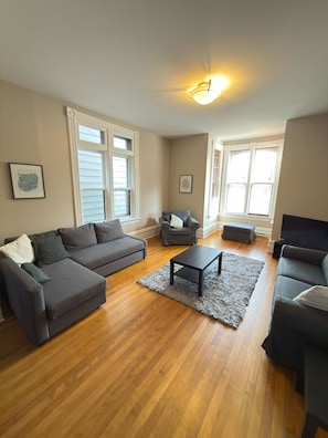 A living room with a grey sectional sofa, a grey armchair, a small ottoman, a coffee table, and a flat-screen TV. - Sunny and airy downtown apartment in Hull Gatineau (Gatineau)