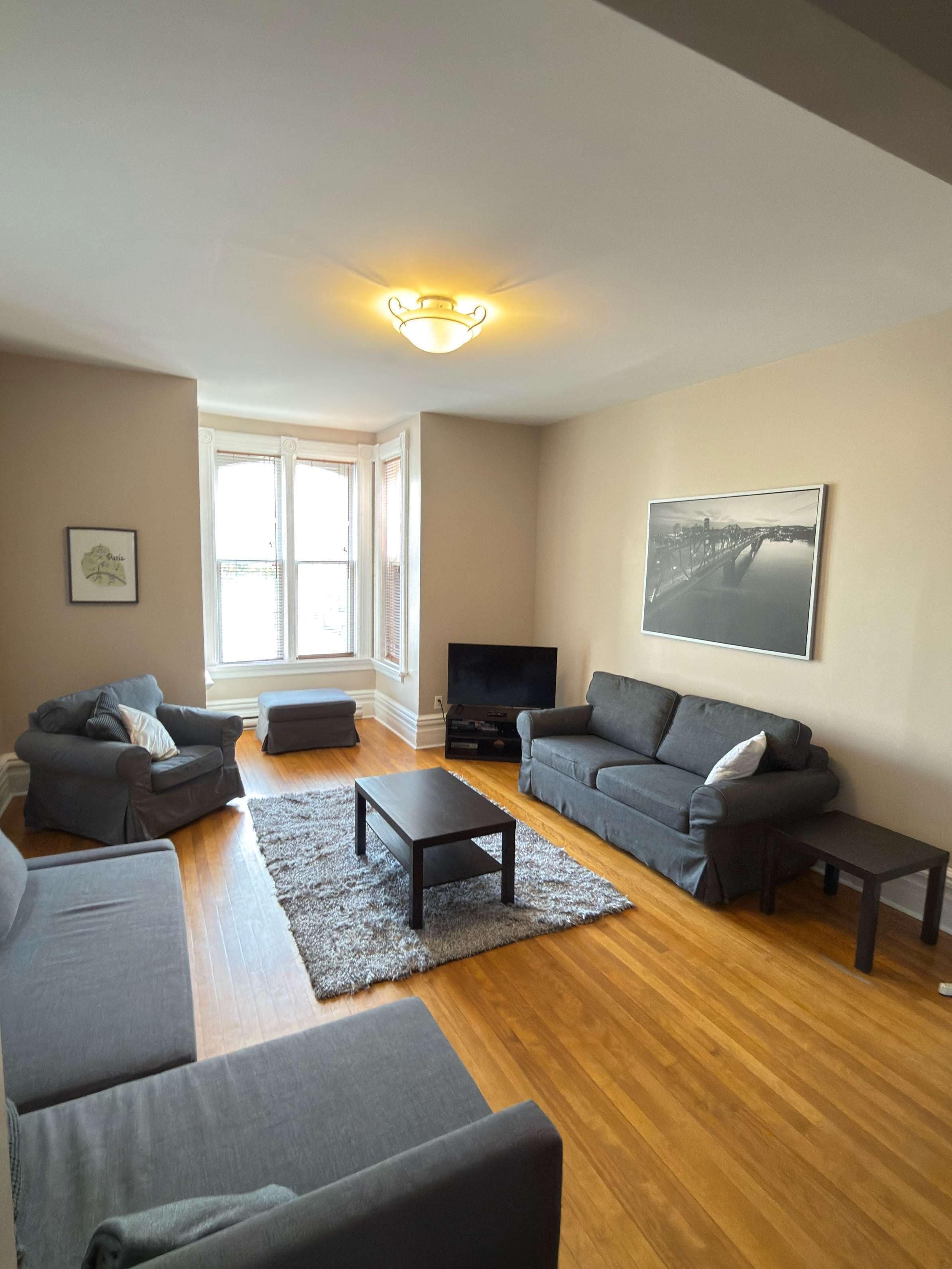 A living room with two grey sofas, a coffee table, a TV, and a large window with blinds.
