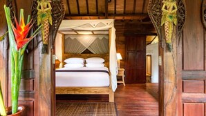 A wooden four-poster bed with white linens, a woven mat at the foot, and a nightstand with a lamp. - Be Bali Hut Farm Stay (Ubud)