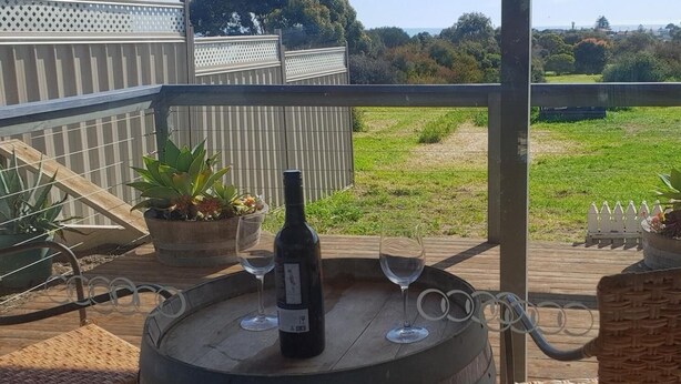 A wooden barrel table with a bottle of wine and two glasses on a balcony with a view of a grassy field and a wooden fence. - Trading Places (Penneshaw)