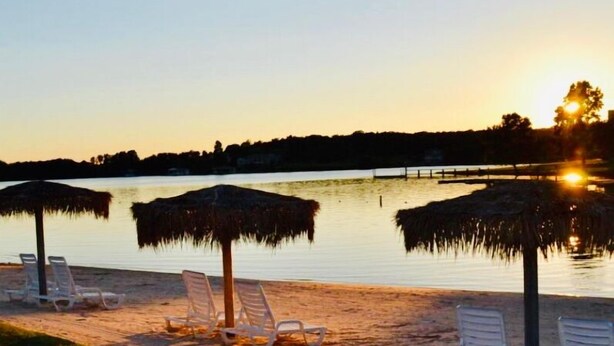 Une plage avec des chaises longues et des parasols, un lac paisible et un coucher de soleil en arrière-plan.