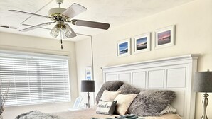 Une chambre à coucher comprenant un lit, des tables de chevet, une commode, une chaise, un ventilateur de plafond et une fenêtre avec des stores.