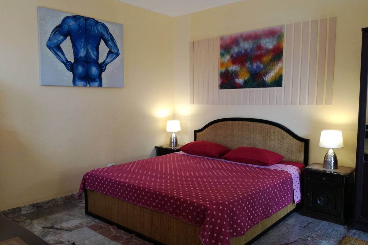 Une chambre à coucher bien rangée, avec un lit, deux tables de chevet dotées de lampes, une couvre-lit à motifs rouges et blancs, et deux œuvres d’art murales.