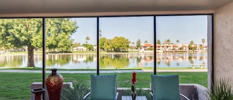 A balcony with two green chairs, a small table, and a vase with red flowers. A large window offers a view of a lake and buildings in the distance.