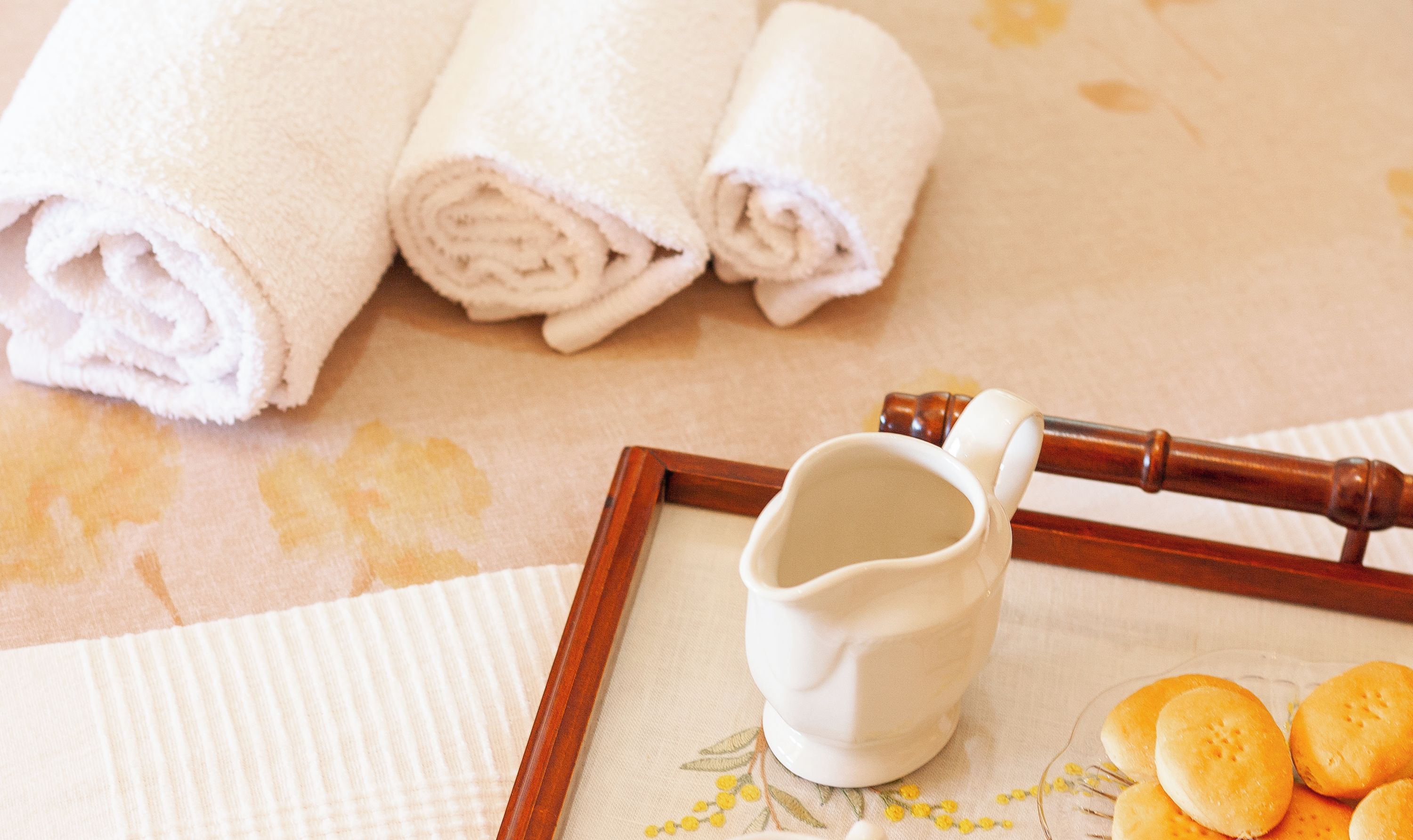 A neatly folded white towel, a white ceramic pitcher, a white cup, and a plate with bread rolls on a table with a floral tablecloth.