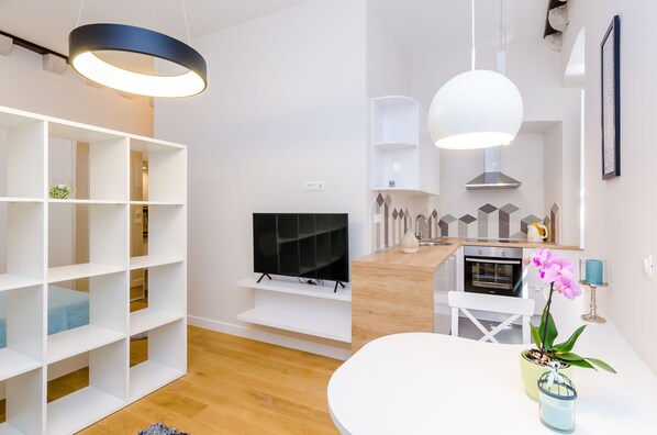 Un salon moderne avec un téléviseur, des étagères blanches, une table à manger et un coin cuisine équipé d’un four.