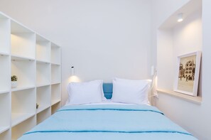 A bedroom with a bed, white walls, a blue headboard, a shelf with a plant, and a framed picture on the wall.