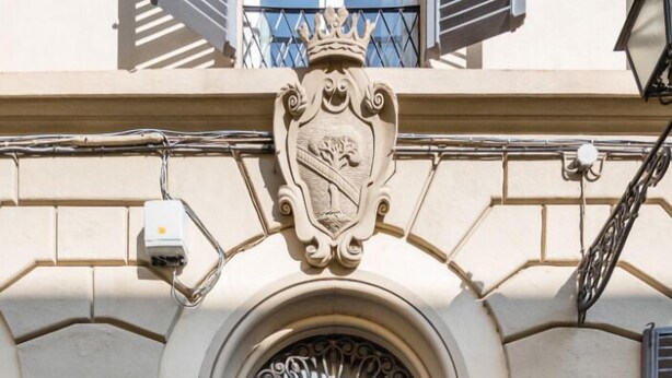 Una puerta doble de madera con un escudo decorativo encima, en un edificio color beige con una placa de número en la pared.