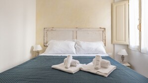 Una cama con sábanas blancas, dos toallas enrolladas y una colcha azul.
