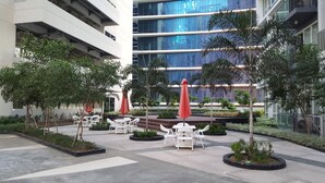 Un espace extérieur moderne avec des chaises blanches et des parasols rouges, entouré de verdure et d’un bâtiment aux fenêtres en verre réfléchissant.